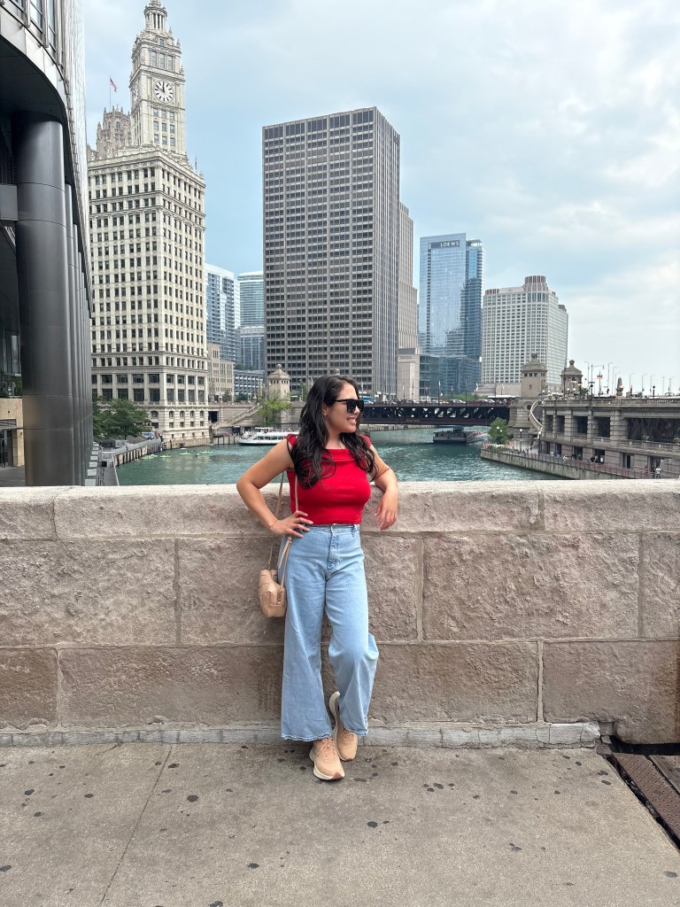 Uma mulher com blusa vermelha e calças jeans largas posa ao lado de um canal em Chicago, com arranha-céus ao fundo.