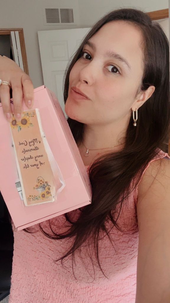 Pessoa segurando uma caixa rosa com um cartão decorativo em destaque, sorrindo para a câmera.