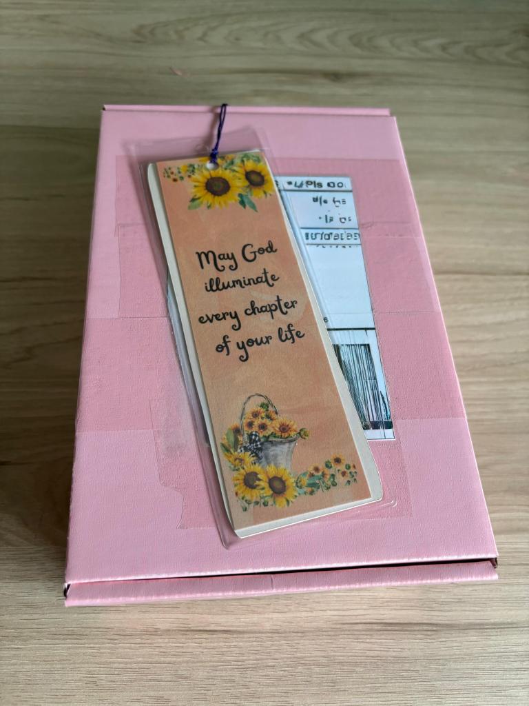 Um marcador de página com flores e a frase 'Que Deus ilumine cada capítulo de sua vida' sobre uma caixa rosa.